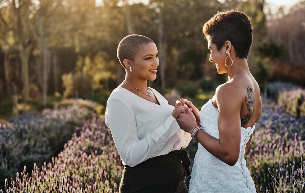 Two brides holding hands and smiling at each other outdoors.